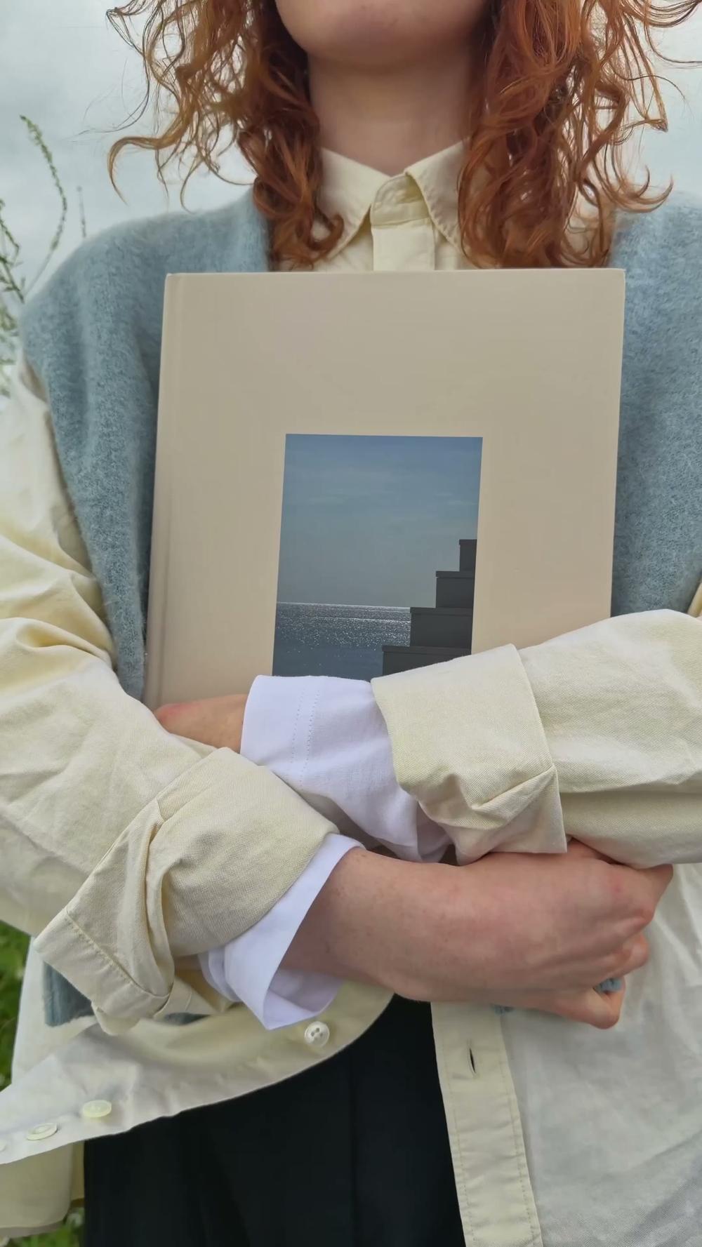 Someone holds a cream foto foto hardcover against their chest, the cover photo showing concrete steps running down to the sea.